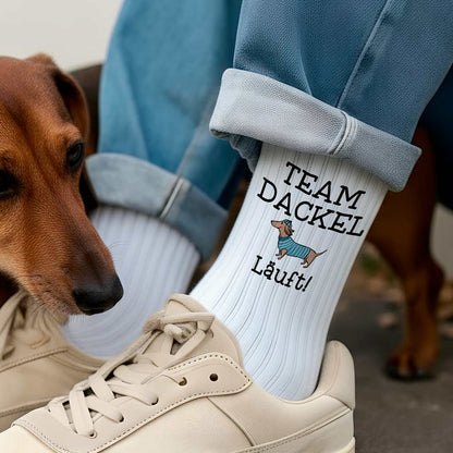 Eine Person trägt weiße Retro-Tennissocken mit einem Dackel-Motiv und dem Spruch 'Team Dackel läuft'.