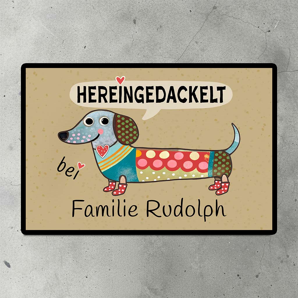 Eine personalisierte Fußmatte mit einem Dackel-Motiv und dem Spruch 'Hereingedackelt' auf dem Steinboden vor einer Haustür.