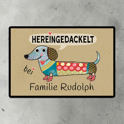 Eine personalisierte Fußmatte mit einem Dackel-Motiv und dem Spruch 'Hereingedackelt' auf dem Steinboden vor einer Haustür.